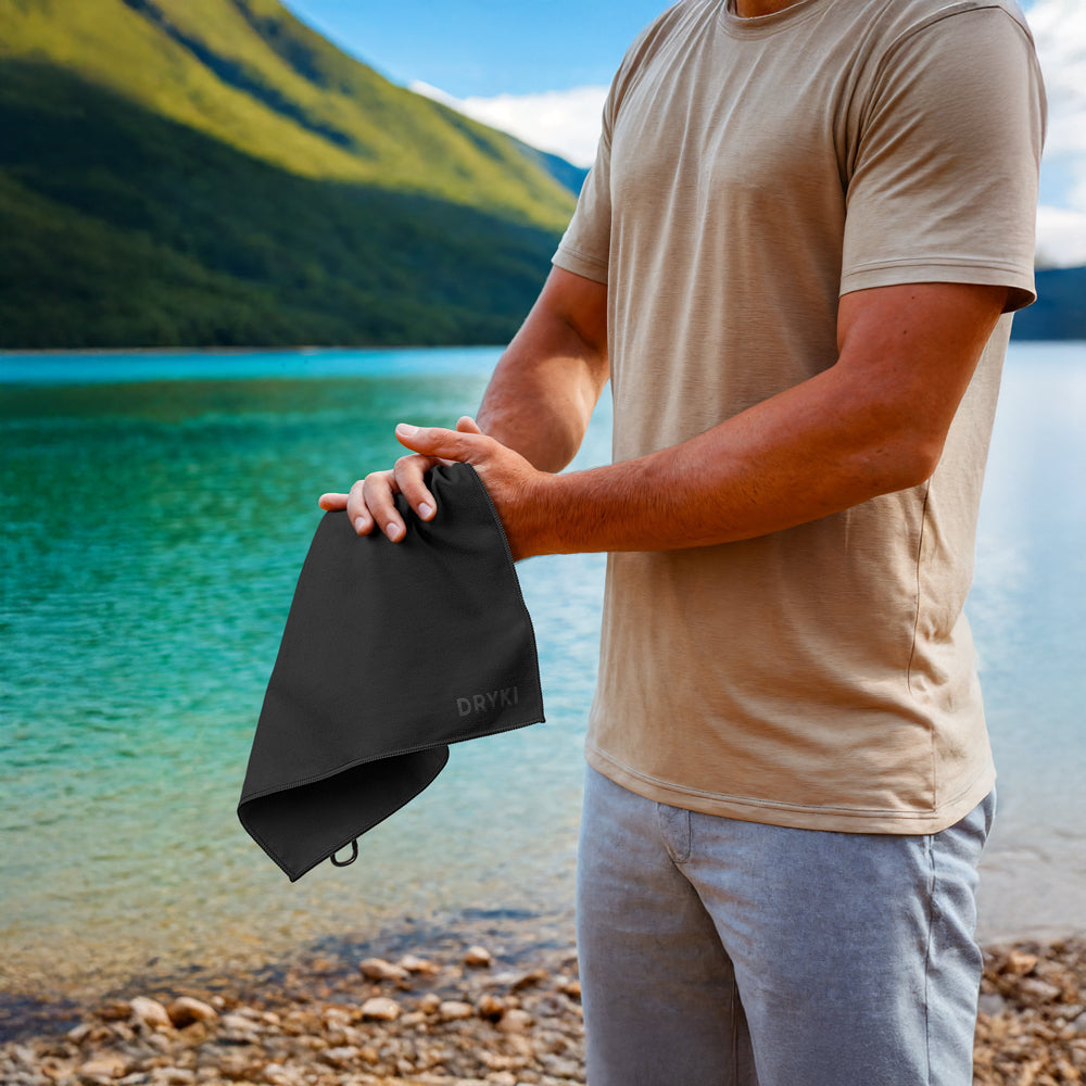 Person holding a black DRYK towel by a lake with mountains in the background, wiping hands with a DRYKI Pocket-sized sweat towel (sweat absorbing handkerchief)