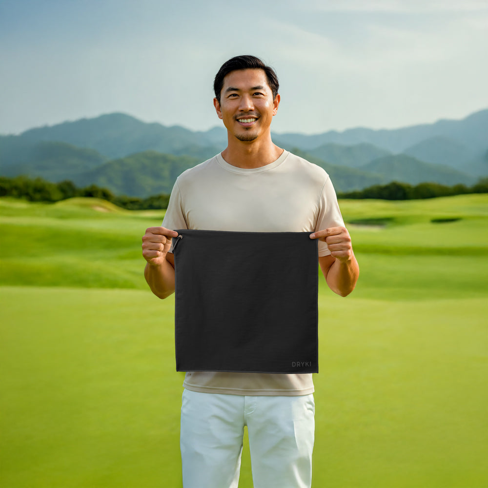 Man holding a black rectangular object on a golf course with mountains in the background, with a DRYKI Pocket-sized sweat towel (sweat absorbing handkerchief)